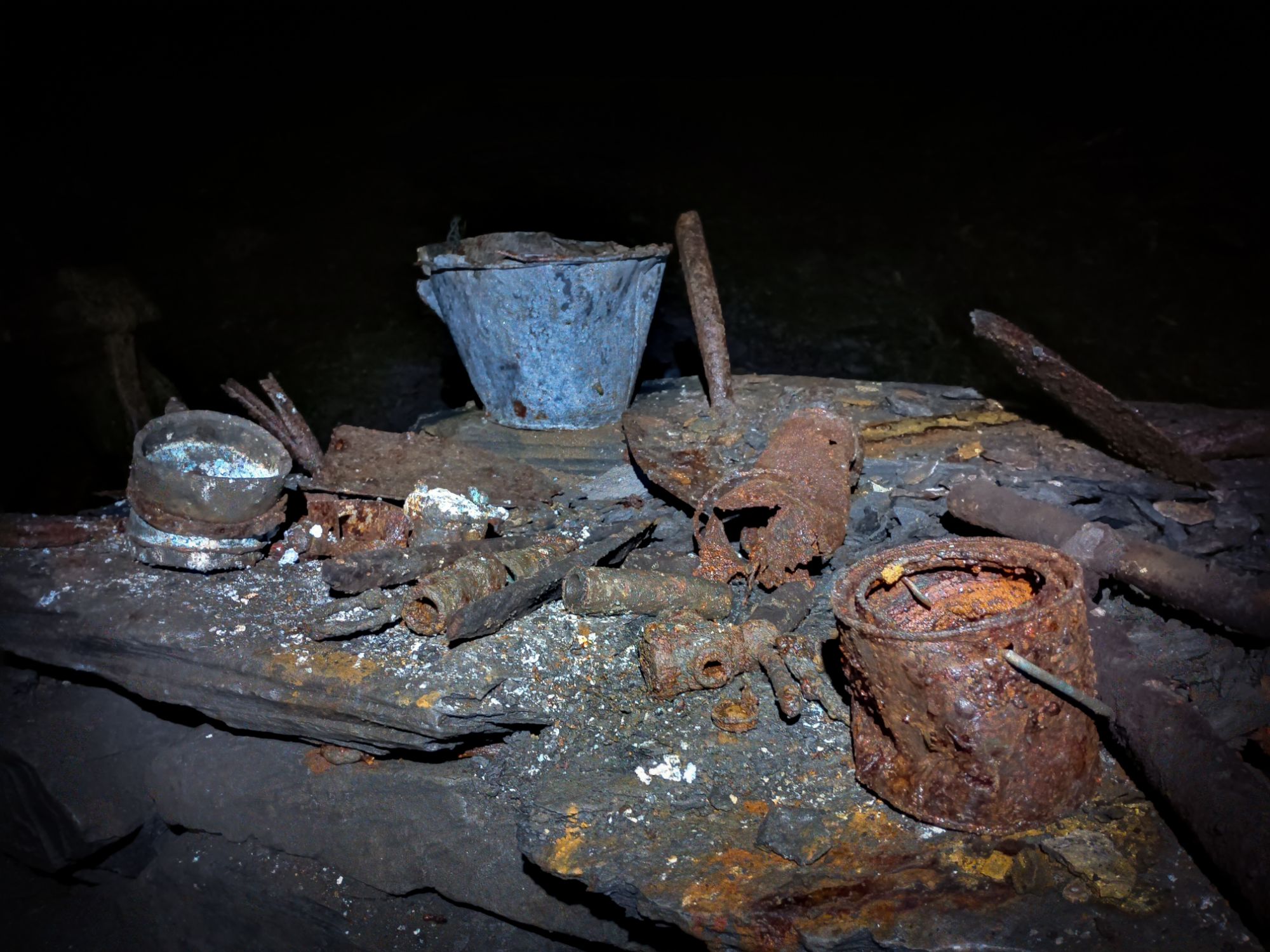 The image titled "Rusting Items Abandoned By Miners In Moel Fferna Slate Mine" shows a collection of old, rusting items left behind by miners in Moel Fferna Slate Mine, North Wales. The items include valves, an old shovel, and rusting paint cans. The scene captures a sense of abandonment and history, with the rust and decay highlighting the passage of time since these items were last used. The setting evokes a sense of nostalgia and intrigue, as it provides a glimpse into the past lives of the miners who once worked in the slate mine.